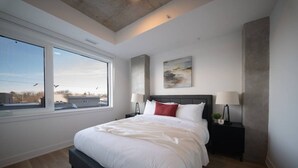 Un dormitorio moderno con una cama grande, vista por una ventana, un cuadro en la pared y una mesita de noche con una lámpara.