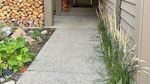 Un passage couvert mène à l’entrée d’une maison, où se trouvent une porte, une pile de bois de chauffage, une plante en pot et une petite citrouille.