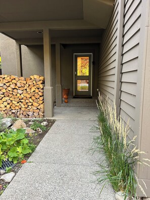 A covered walkway leading to a house entrance with a door, a stack of firewood, a potted plant, and a small pumpkin. - Frenchman’s View charming home near River Run (Ketchum)