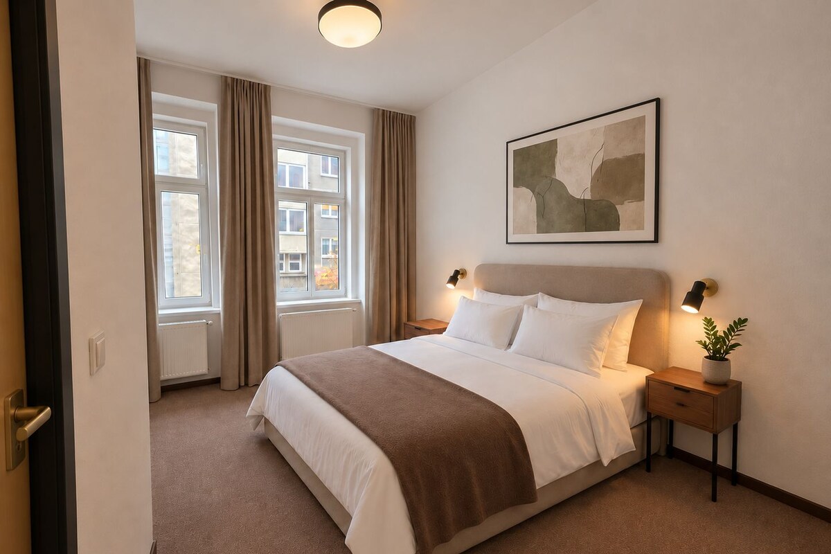 A hotel room with a large bed, a bedside table with a lamp, a window with curtains, and a painting on the wall.