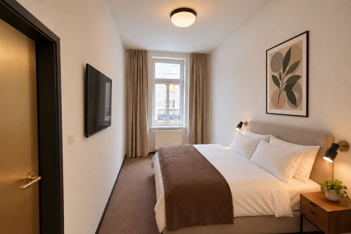 A hotel room with a bed, a bedside lamp, a window with curtains, a wall-mounted TV, and a framed picture.