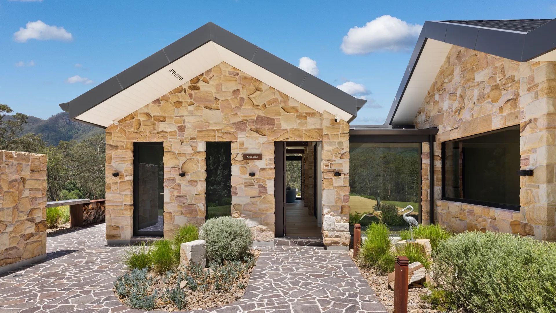A stone house with a modern design, featuring a stone facade, large windows, and a paved entrance area with landscaping.