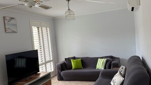 Ein Wohnzimmer mit einem Flachbildfernseher, einem dunklen Sofa mit grün-weiß gestreiften Kissen, einer blauen Decke, einem Holztisch und einem Deckenventilator.