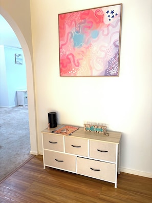 A room with a wooden floor, a white dresser with drawers, a framed abstract painting, and a small table with a magazine and a black object.