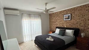 Ein Schlafzimmer mit Bett, Nachttisch, Schreibtisch, Deckenventilator und einem Fenster mit Vorhängen.