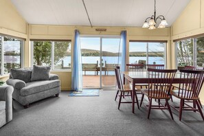 A room with a sofa, dining table, chairs, and large windows offering a view of a lake.