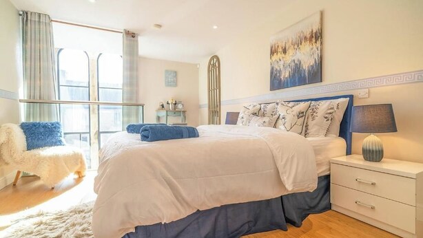 Une chambre à coucher avec un lit, une table de chevet, une lampe, une chaise et une fenêtre avec des rideaux.