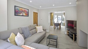 A modern living room with a grey sofa, a small table, a TV, and a dining area with yellow curtains. - Bright 3-Bed House in Watford Near London Links & Studios (Hertfordshire)