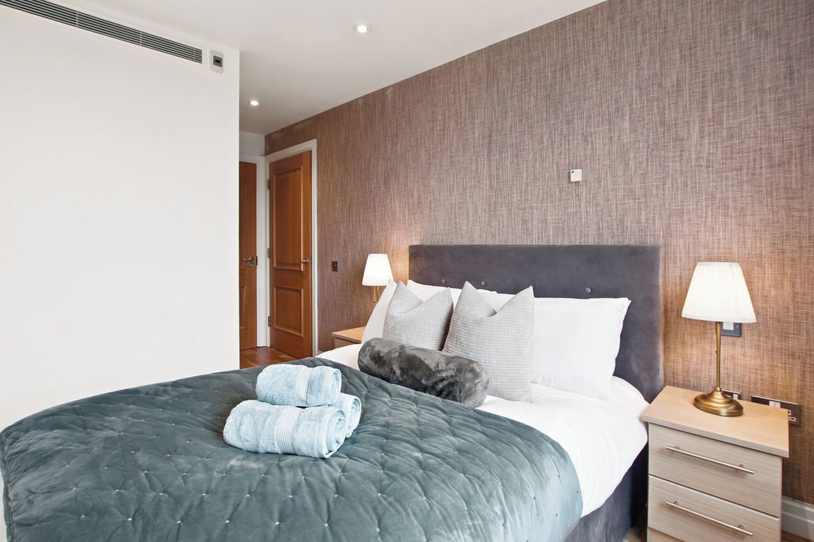A neatly made bed with a grey quilt, two rolled towels, and a grey cat on the bed. There are two bedside lamps, a wooden nightstand, and a door in the background.