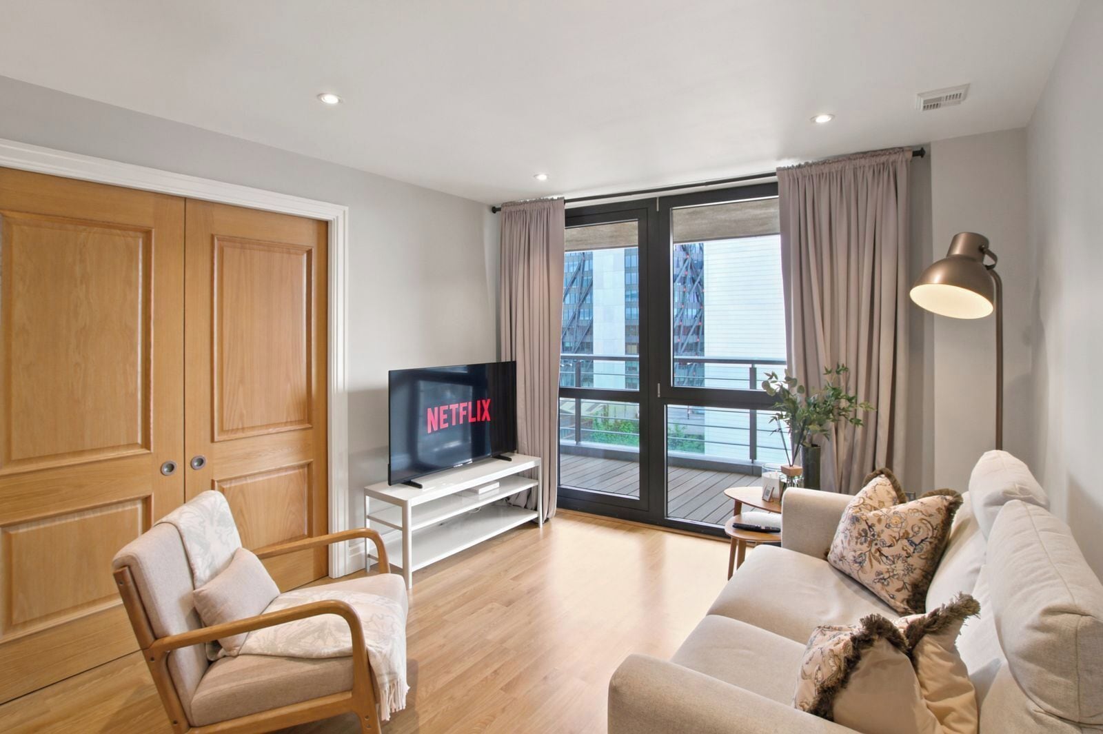 Uma sala de estar moderna com uma TV exibindo 'NETFLIX', um sofá, uma poltrona e uma janela grande com cortinas.