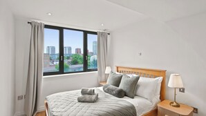 Ein Schlafzimmer mit einem großen Fenster, einem Bett mit grauer Bettwäsche, zwei Nachttischlampen und einem Holznachttisch.