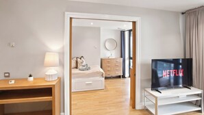 Ein modernes Hotelzimmer mit einem Holztisch, einer Nachttischlampe, einem Bett mit zusammengerollten Handtüchern, einem Schminktisch und Blick durch eine geöffnete Tür.