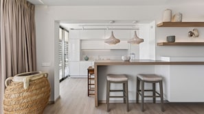 Eine moderne Küche mit weißen Schränken, einer Holzinsel mit Barhocker, Pendelleuchten und einem Korbwäscheschrank.