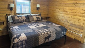 Une chambre dans une cabane en bois, avec un lit, deux lampes fixées au mur, une fenêtre avec des stores et un plafond en bois.
