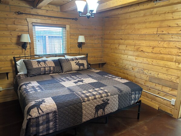 Un dormitorio de cabaña de madera con cama, dos lámparas fijadas a la pared, una ventana con persianas y techo de madera.