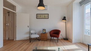 Une pièce avec un plancher en bois, une porte blanche, une œuvre d’art encadrée au mur, une chaise rouge et deux lampadaires.