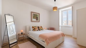 Ein Schlafzimmer mit einem Bett, einem Nachttisch, einer Lampe, einem Spiegel, einem Fenster und einer Pendelleuchte.