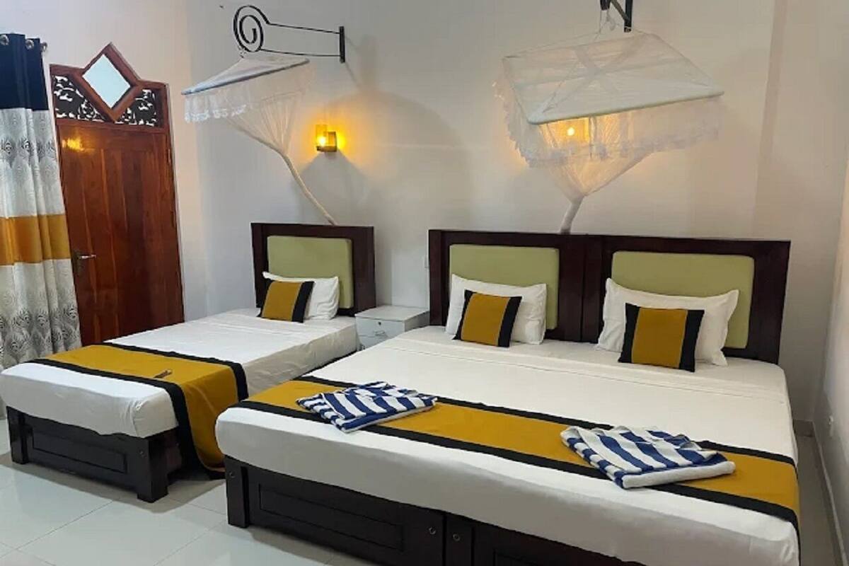 Habitación de hotel con dos camas, cada una con colcha amarilla y ropa de cama blanca, una puerta de madera y una lámpara fijada a la pared.