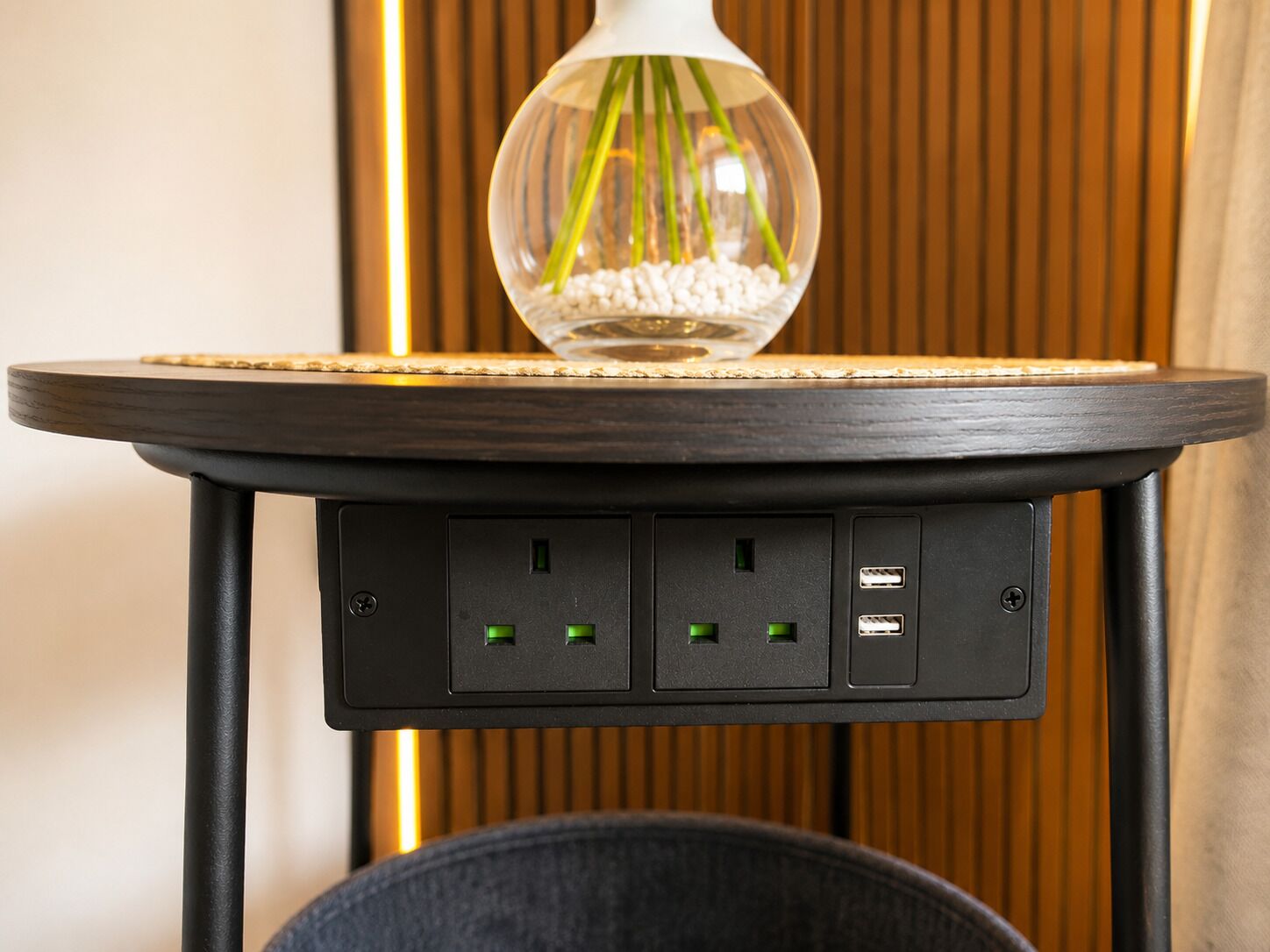 A table with a glass vase containing green plants and white pebbles, a round wooden top, and a black base with electrical outlets.