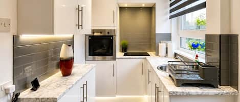 Una cocina moderna con muebles blancos, horno empotrado, lavamanos y ventana con persianas.