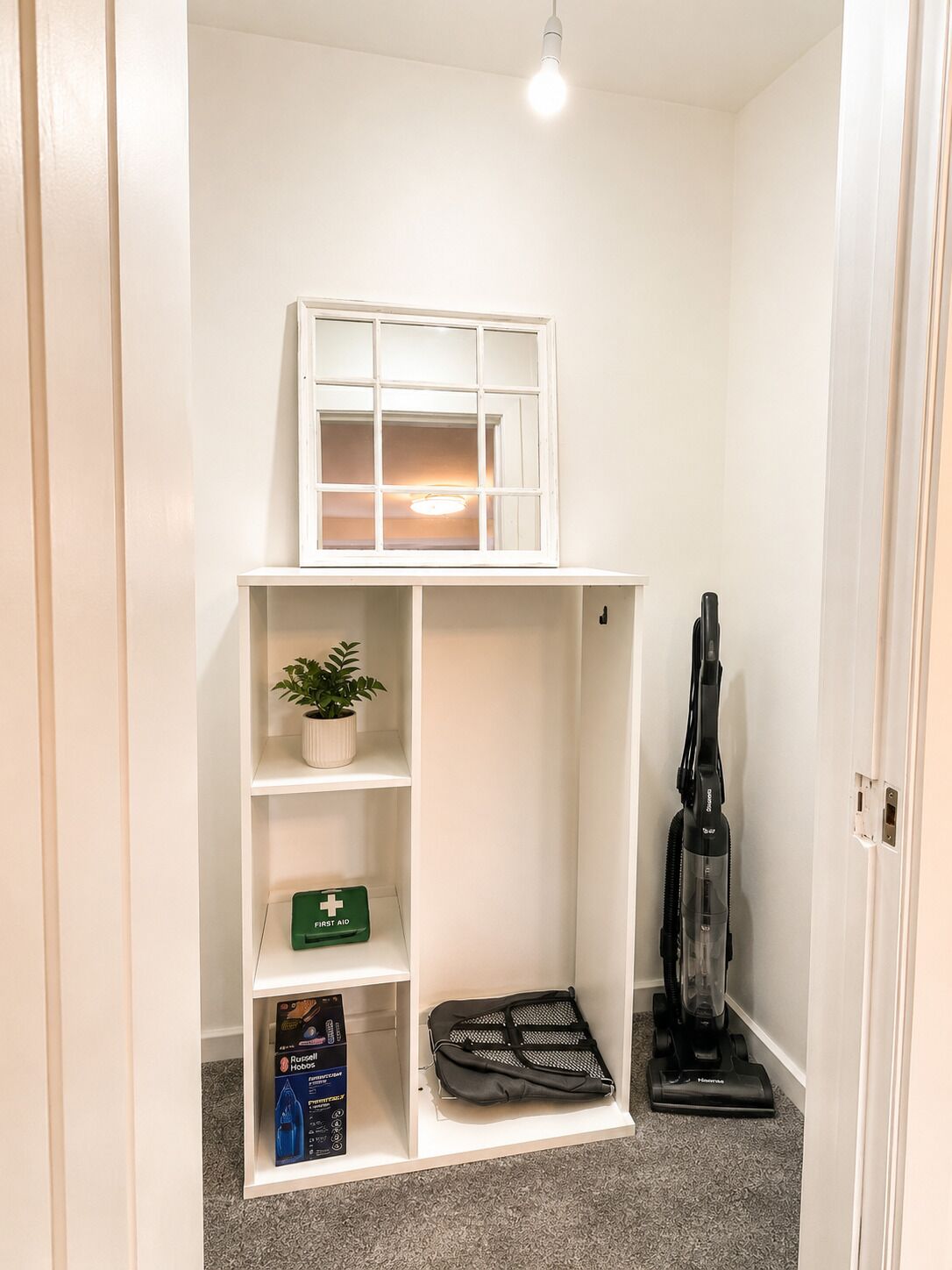 Una habitación pequeña con un estante blanco, una ventana diminuta, un aspirador y un botiquín de primeros auxilios.