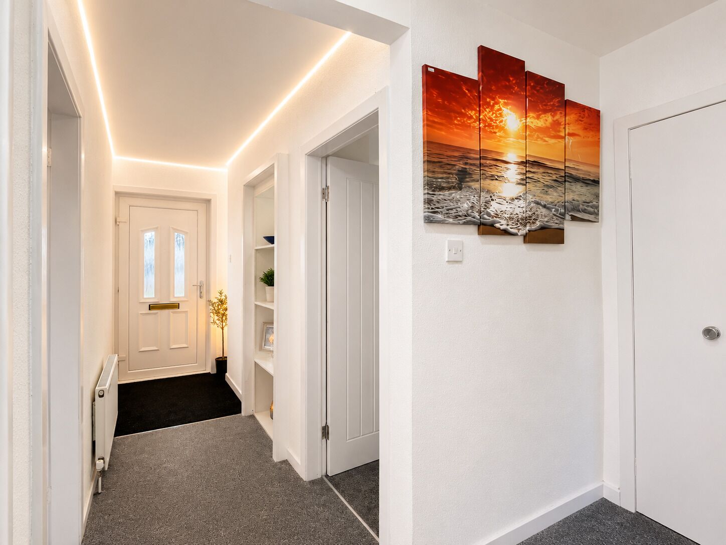 A hallway with a white door, a shelf with a potted plant, and a wall-mounted artwork depicting a sunset over the ocean.
