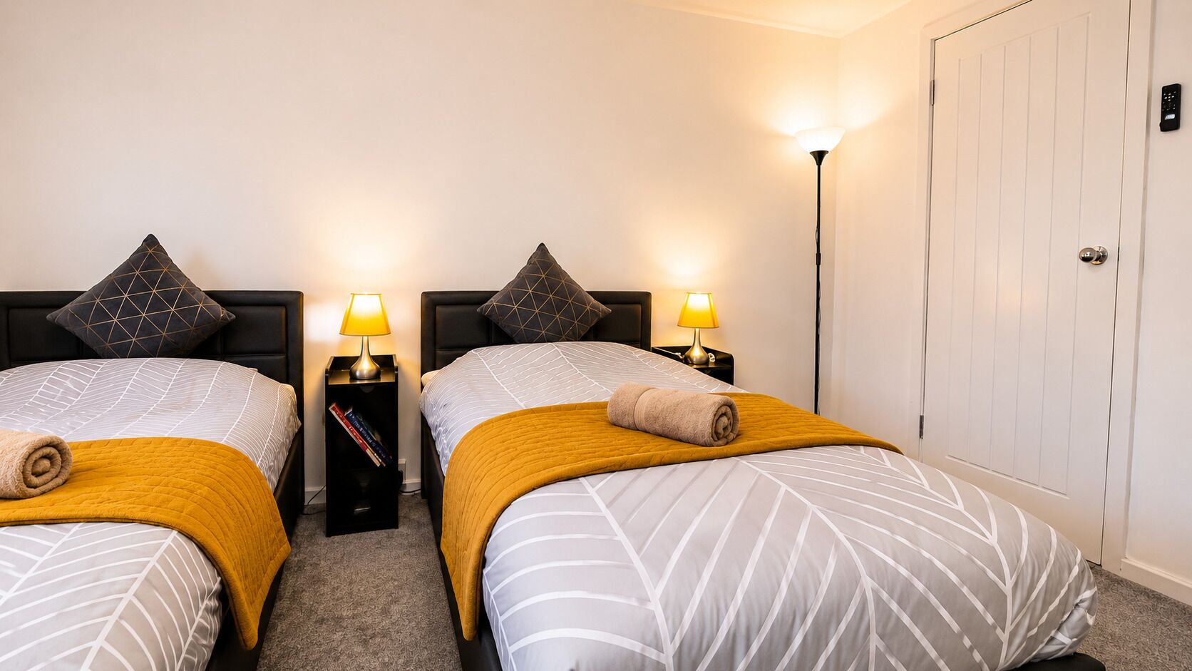 A hotel room with two beds, each with a headboard, a bedside table with a lamp, and a door with a handle.