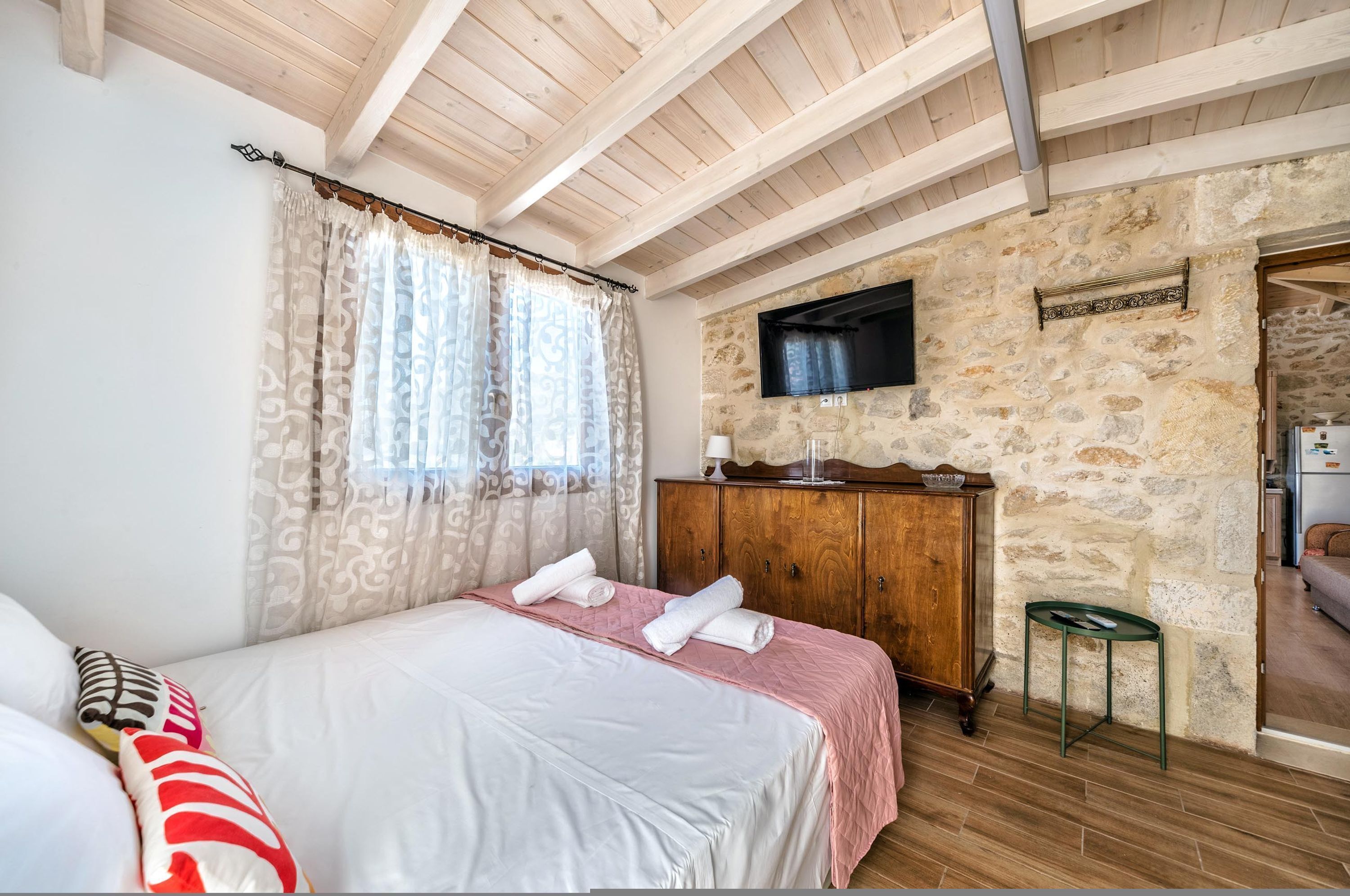 A bedroom with a bed, wooden wardrobe, stone wall, and a television mounted on the wall.
