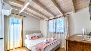 Une chambre avec un plafond en bois, un lit recouvert d’une couette rose, deux serviettes enroulées et une commode en bois surmontée d’un bol en verre.