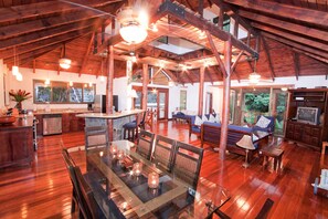 A spacious interior with a high ceiling, wooden beams, a dining area with a glass table, and a living area with a sofa and TV. - Charming 3-bedroom house with AC in beautiful Quepos (Quepos)