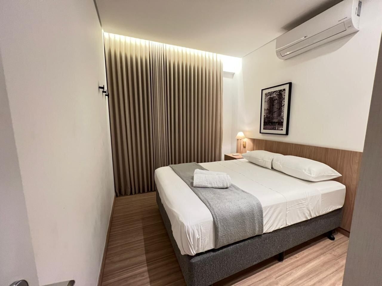 Habitación de hotel con cama, lámpara de noche, aire acondicionado de pared y un cuadro enmarcado.