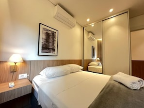 Habitación de hotel con cama, lámpara de noche, equipo de aire acondicionado, un cuadro enmarcado, un espejo y una toalla sobre la cama.