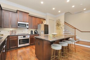 A modern kitchen with wooden cabinets, stainless steel appliances, a central island with bar stools, and a staircase leading to another level. - Spacious and Lux 2 BR With Game Room & Hot Tub + Gated Community (Houston)
