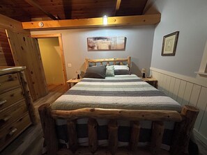 A wooden-framed bed with a striped bedspread, a nightstand with a lamp, and a painting on the wall. - Merry Grinchmas! Theme getaway, limited availability!! Free photo ops! (Embden)