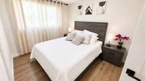 Ein ordentlich bezogenes Bett mit weißer Bettdecke und gemusterten Kissen, ein dunkles Kopfteil, ein Nachttisch mit Lampe und ein kleiner Topfpflanze.
