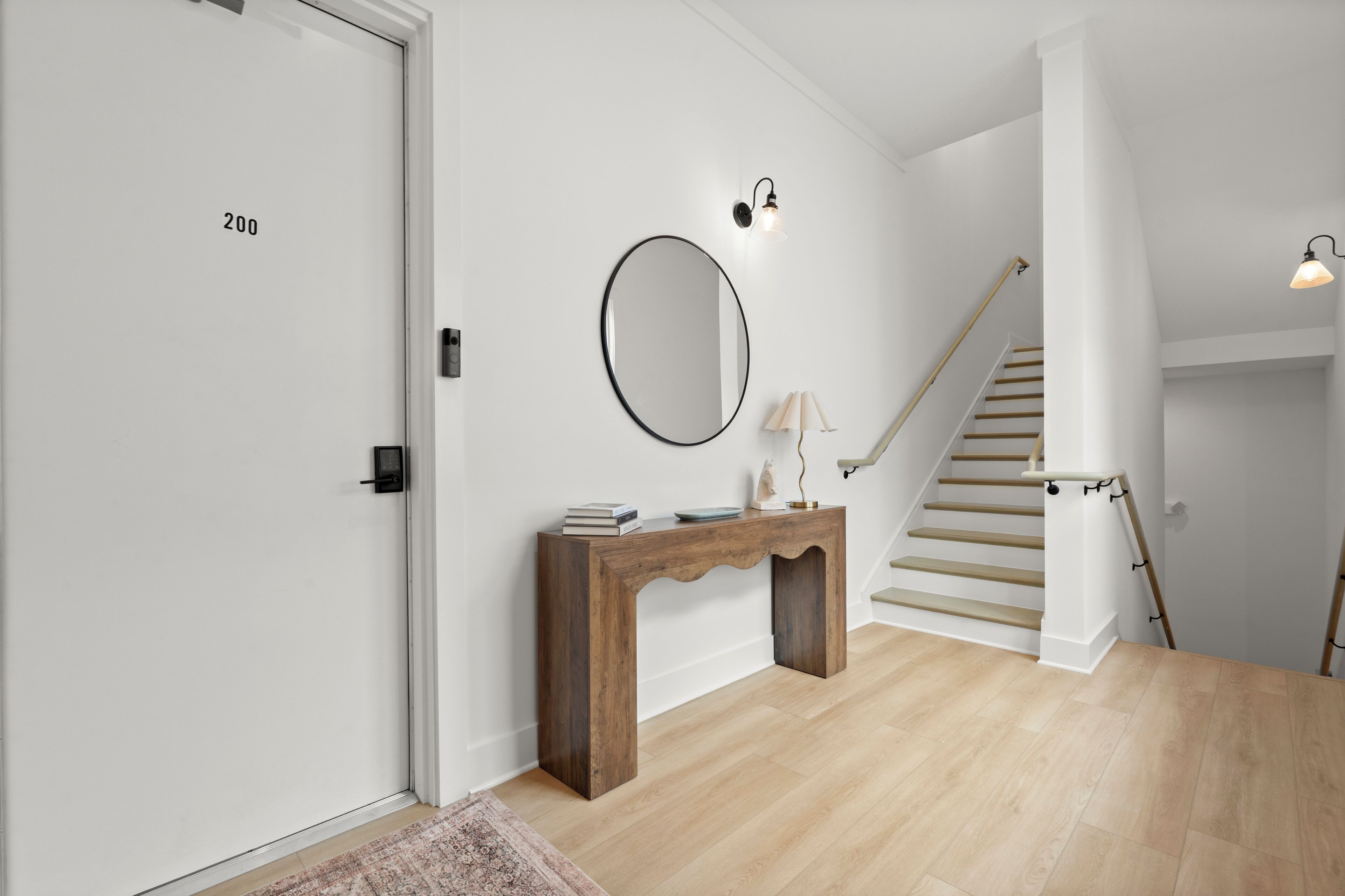A hallway with a wooden console table, a round mirror, a staircase, and a door labeled '200'.