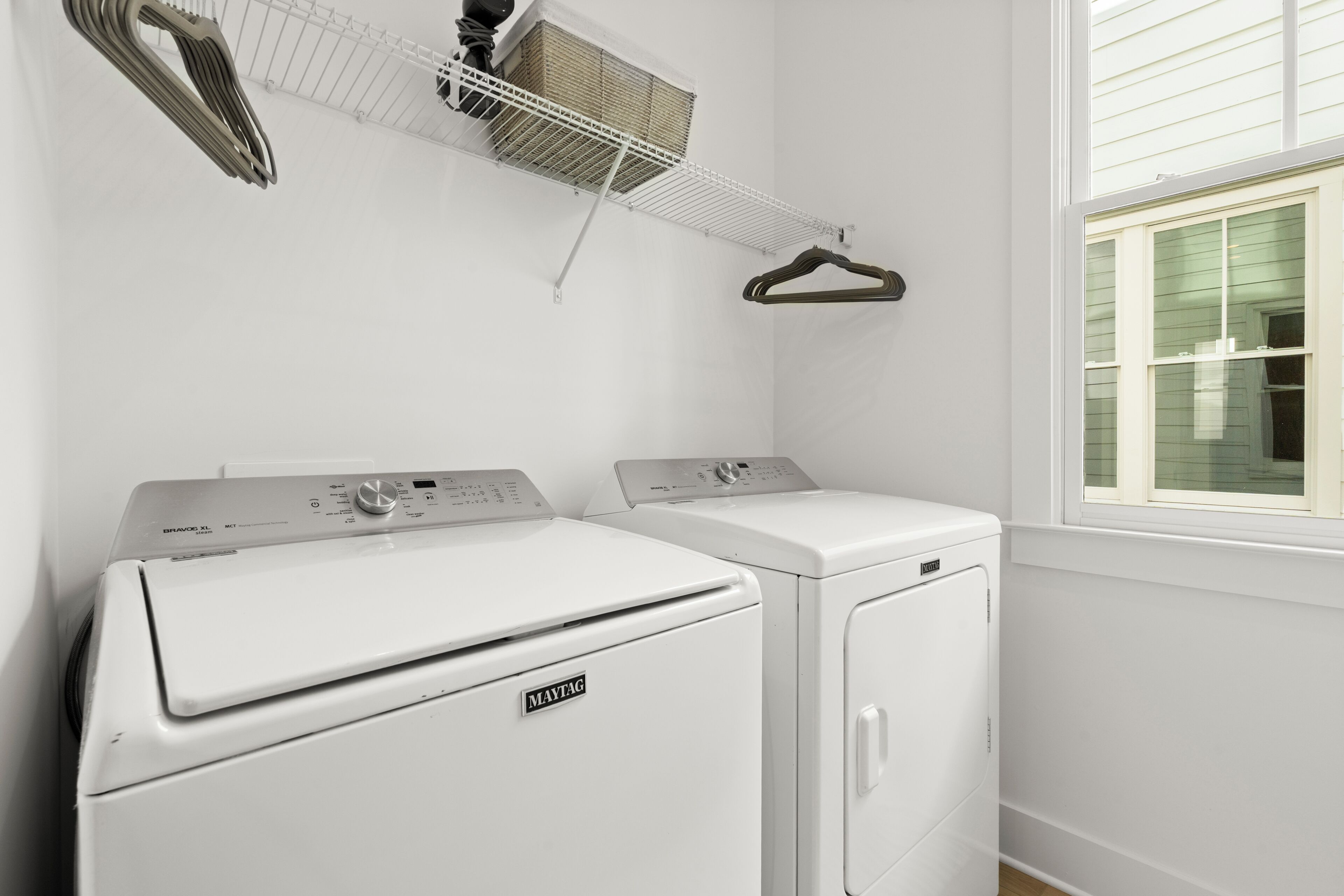 Un área de lavandería con dos secadoras Maytag, un estante blanco con cestos y una ventana con persianas.