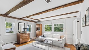 A spacious living room with a white sofa, wooden dresser, and large windows offering a view of greenery. - The School Street House • Dock Square Village (Kennebunkport)