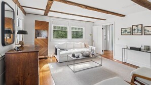 A spacious living room with a white sofa, wooden beams, large windows, and a modern coffee table. - The School Street House • Dock Square Village (Kennebunkport)