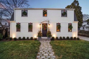 Une maison à deux étages avec un porche à l’avant, un sapin de Noël et des guirlandes aux fenêtres.