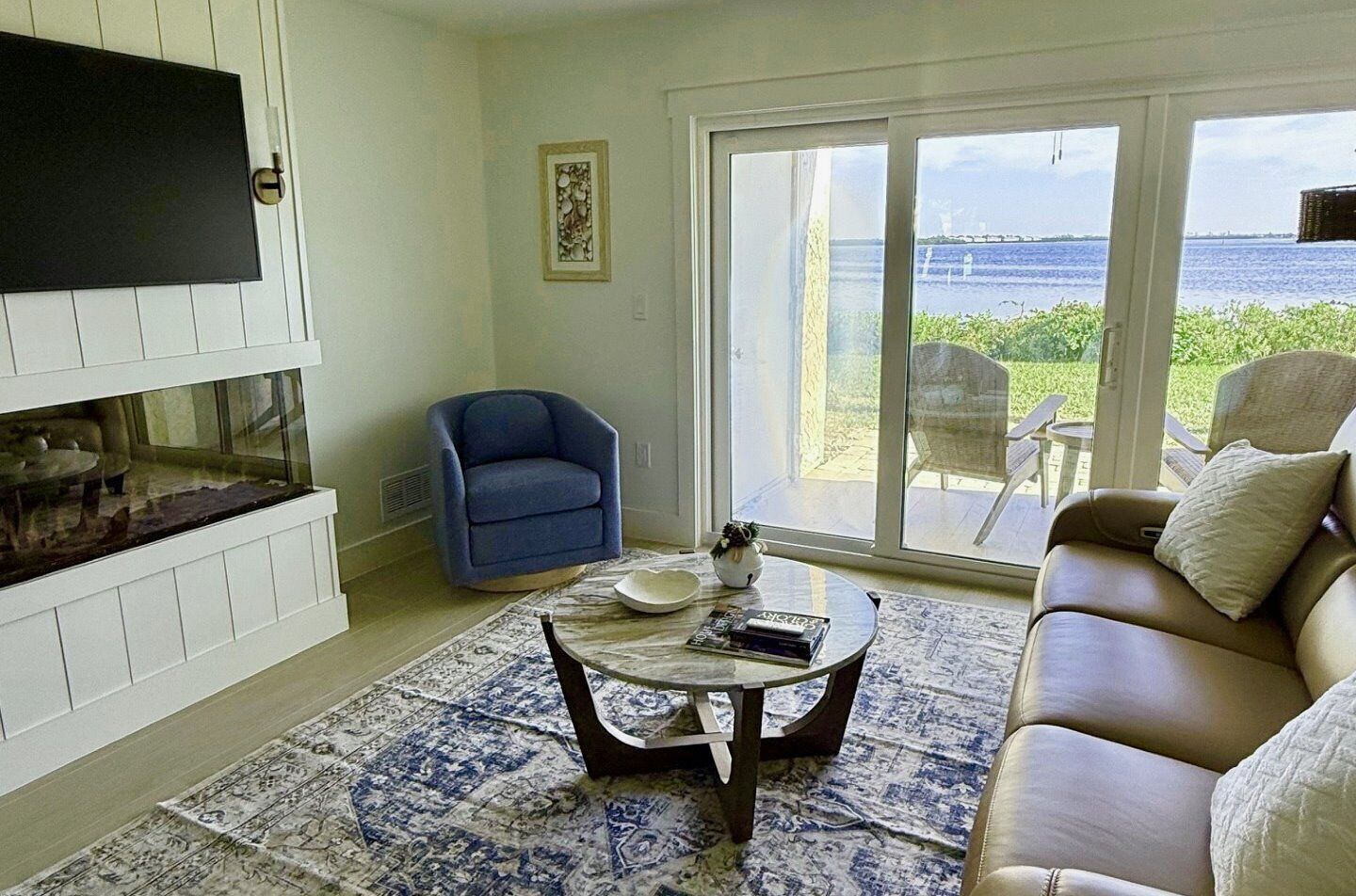 Un salón con televisor, un sillón azul, un sofá y vista al mar a través de amplias ventanas.
