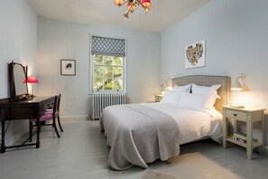 Ein Schlafzimmer mit Bett, Nachttischen, Schreibtisch, Stuhl, Heizkörper, Fenster mit gemustertem Vorhang und einem Wandbild.
