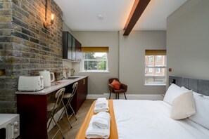 A hotel room with a brick wall, a bed, a desk with a chair, a window, and a view of a building. - Brixton Lofts - Deluxe Studio (No.4) (Bristol)