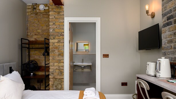 Een hotelkamer met een bed, een kleintje tafeltje met handdoeken, een aan de muur bevestigde televisie, een bakstenen muur en een deur naar een badkamer.