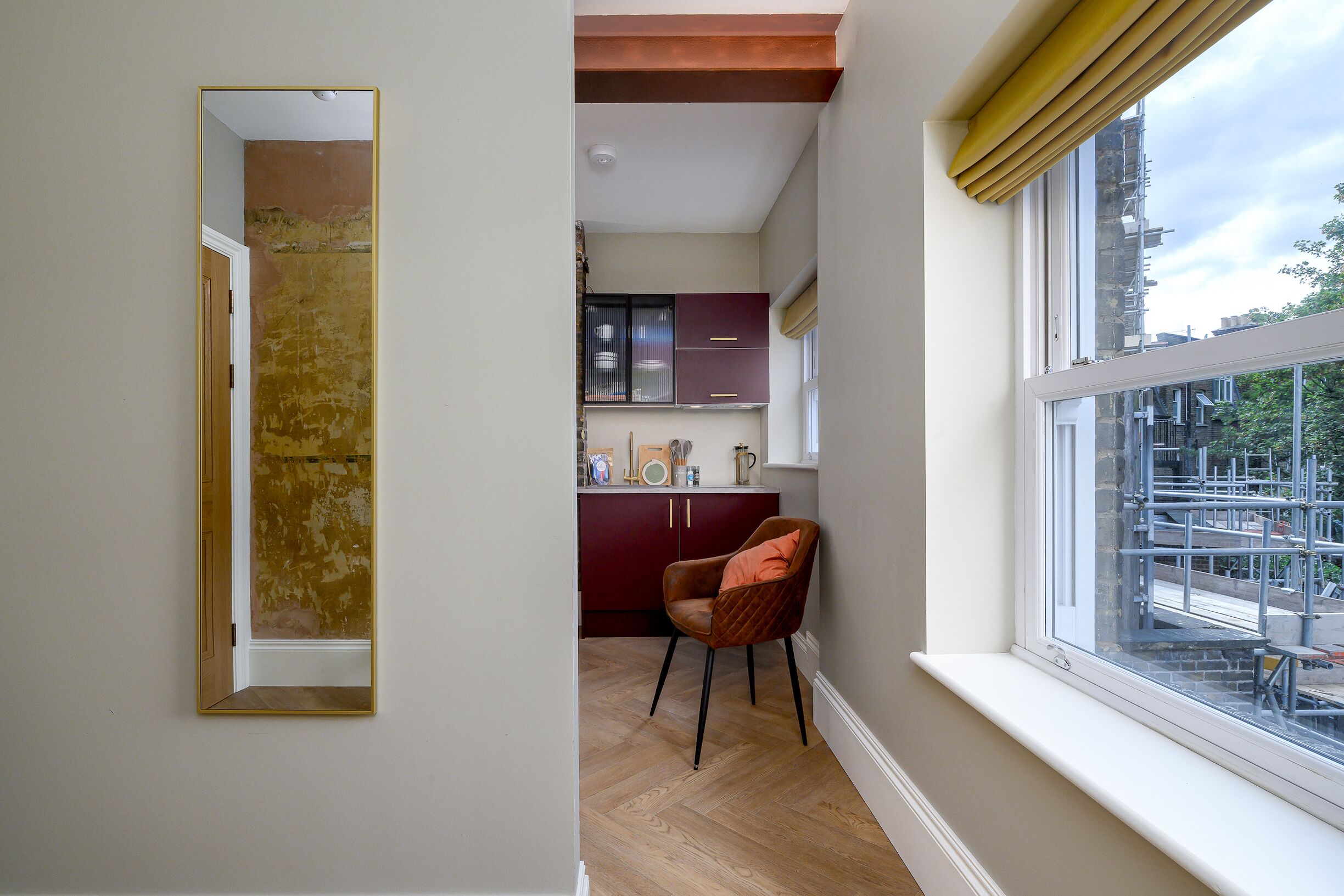 A narrow hallway with a mirror, a red chair, and a window with a view of construction outside.