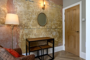A room with a distressed wall, a round mirror, a wooden door, a floor lamp, and a small wooden table. - Brixton Lofts - Deluxe Studio (No.4) (Bristol)