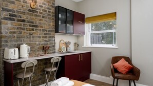 Een compacte kamer met een bakstenen muur, een kitchenette, een bed met witte lakens, een bruine leren fauteuil en een raam met uitzicht.