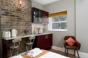 A compact room with a brick wall, a kitchenette, a bed with white linens, a brown leather chair, and a window with a view. - Brixton Lofts - Deluxe Studio (No.4) (Bristol)