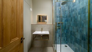 Een badkamer met een houten deur, een wit wastafelmeubel met gouden rand, een spiegel boven de wastafel en een douche met blauwe tegels.