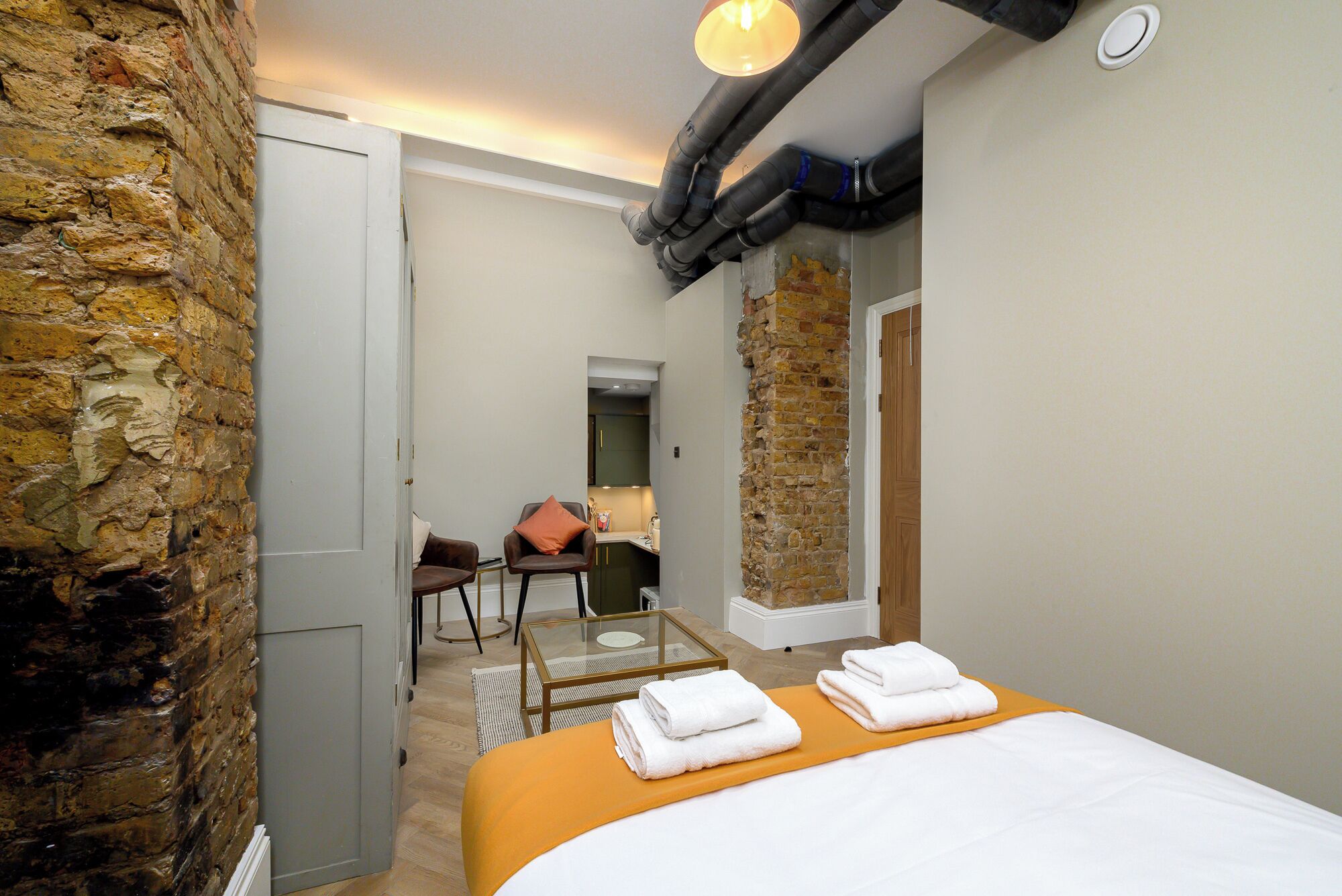 Habitación de hotel con cama, una mesita de vidrio, una silla, pared de piedra y un baño visible al fondo.